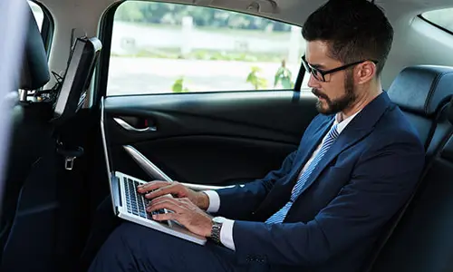 Business professional working on a laptop while riding in the back seat of a private vehicle, representing corporate transportation services.